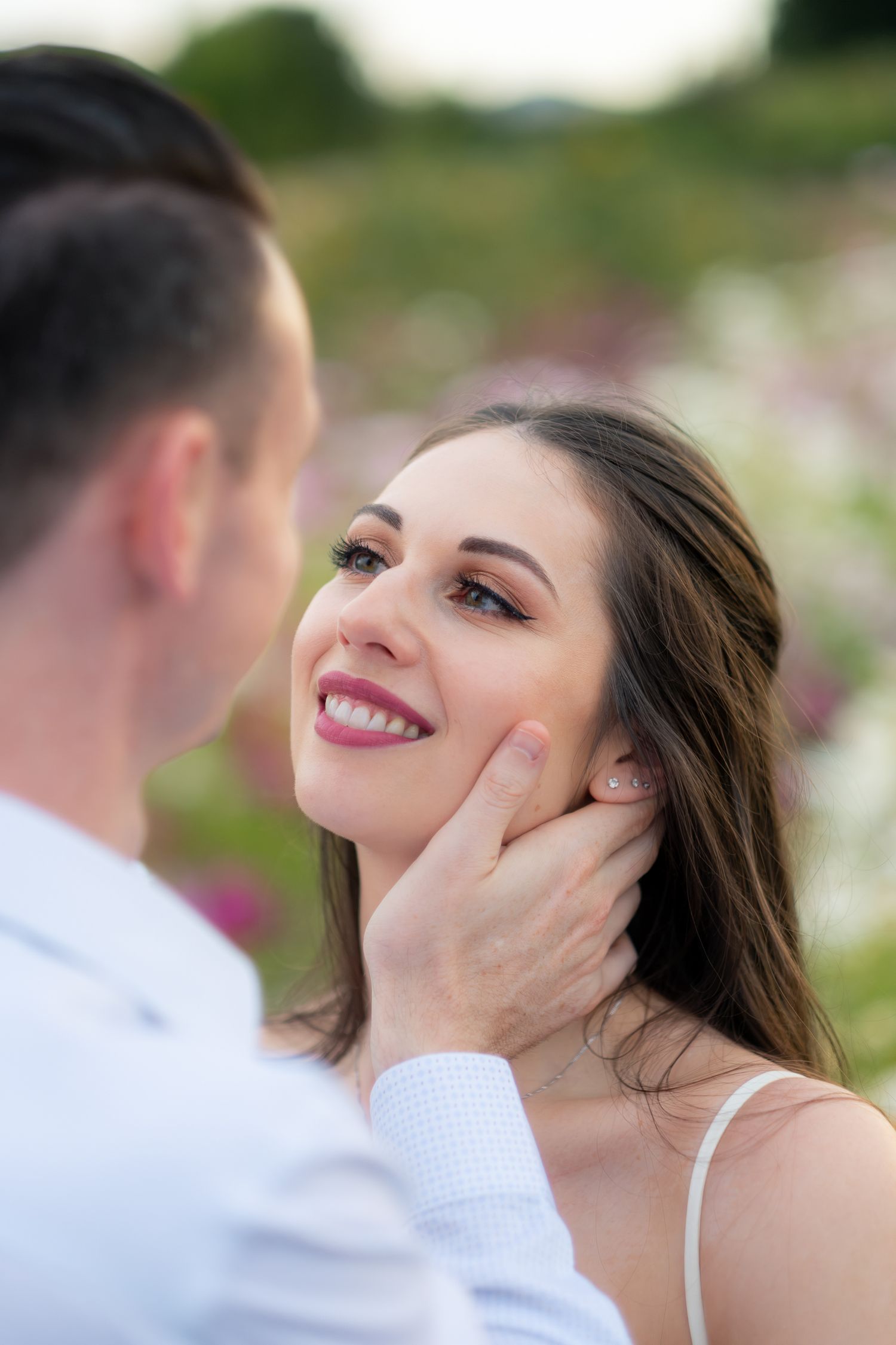 Groom tenderly cups smiling bride's face with colorful flower garden softly blurred in the background.