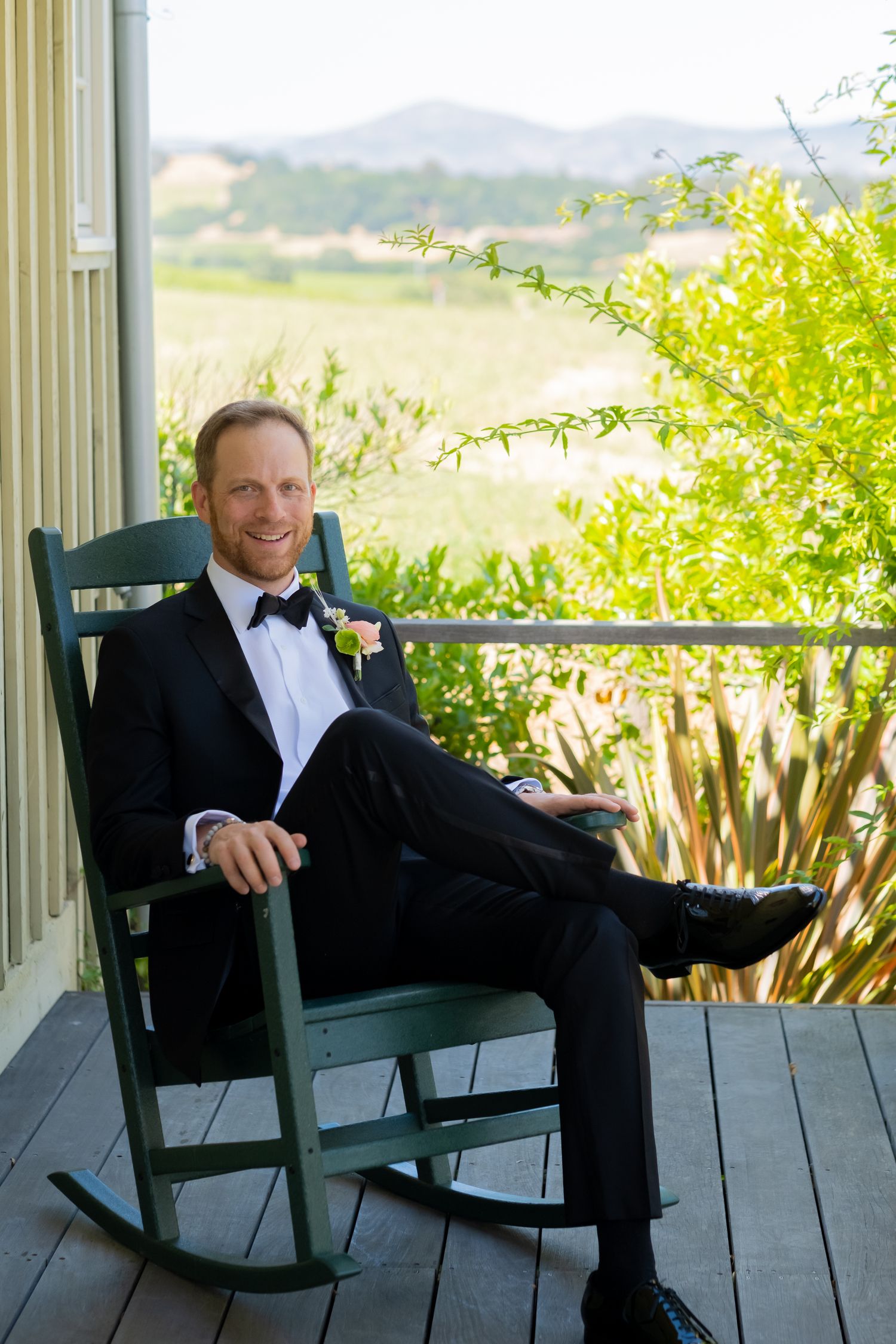 Groom in tuxedo relaxing in green rocking chair on porch with scenic vineyard hills in background.