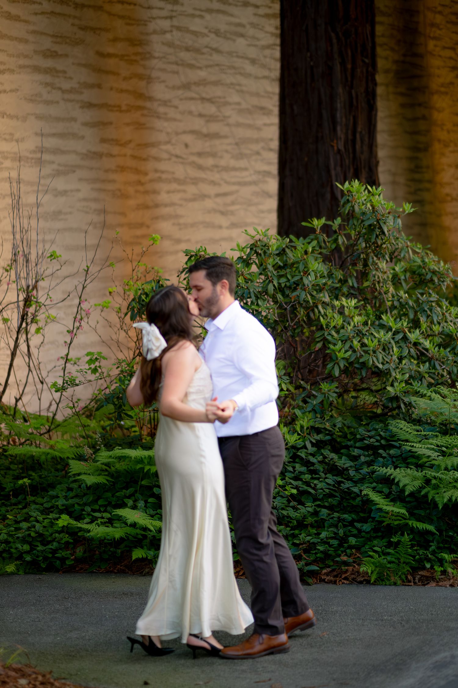 A couple shares a romantic kiss outdoors, bride in white gown with floral crown, groom in white shirt and brown pants.