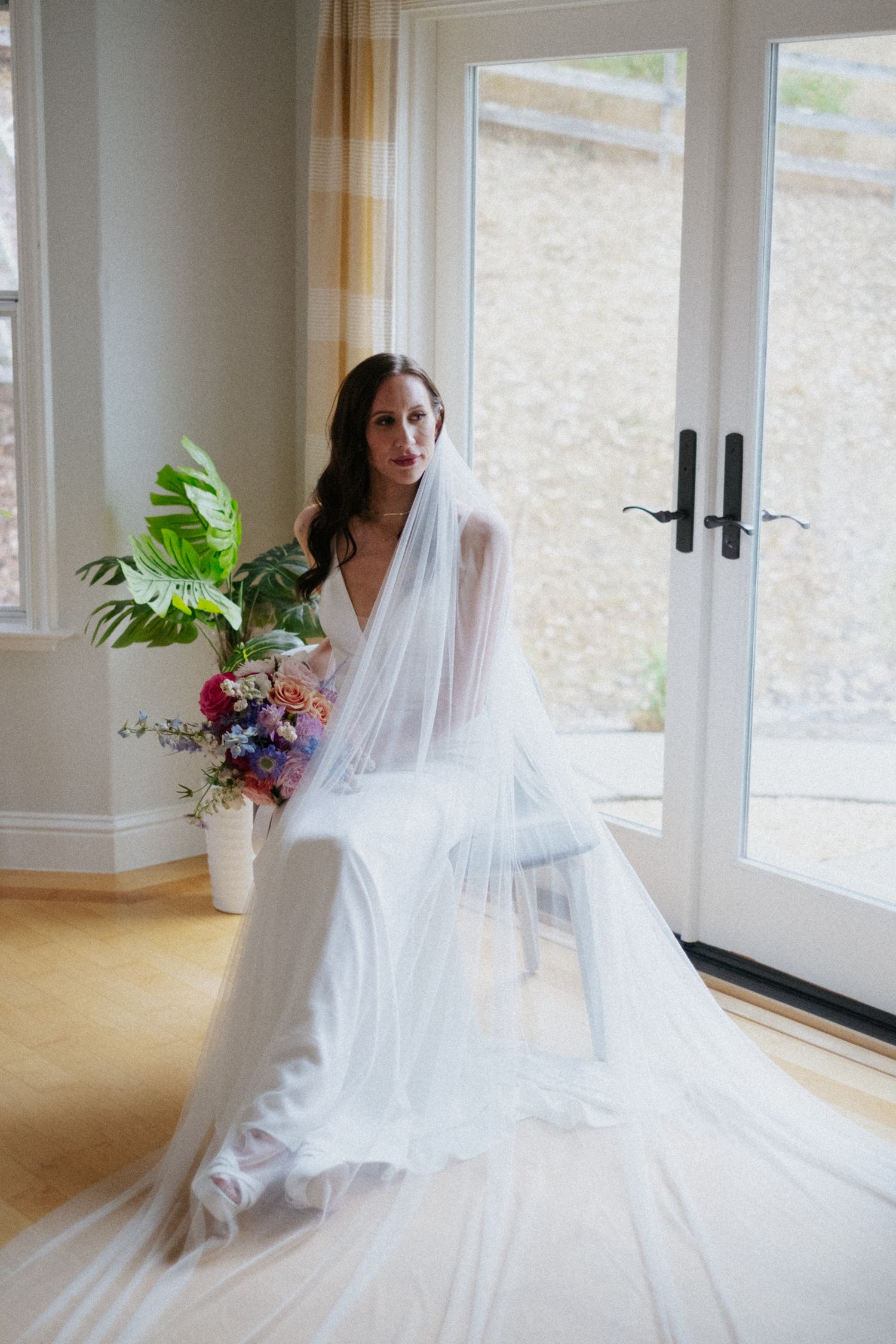 Bride in white wedding dress and long veil seated by French doors holding colorful bridal bouquet.