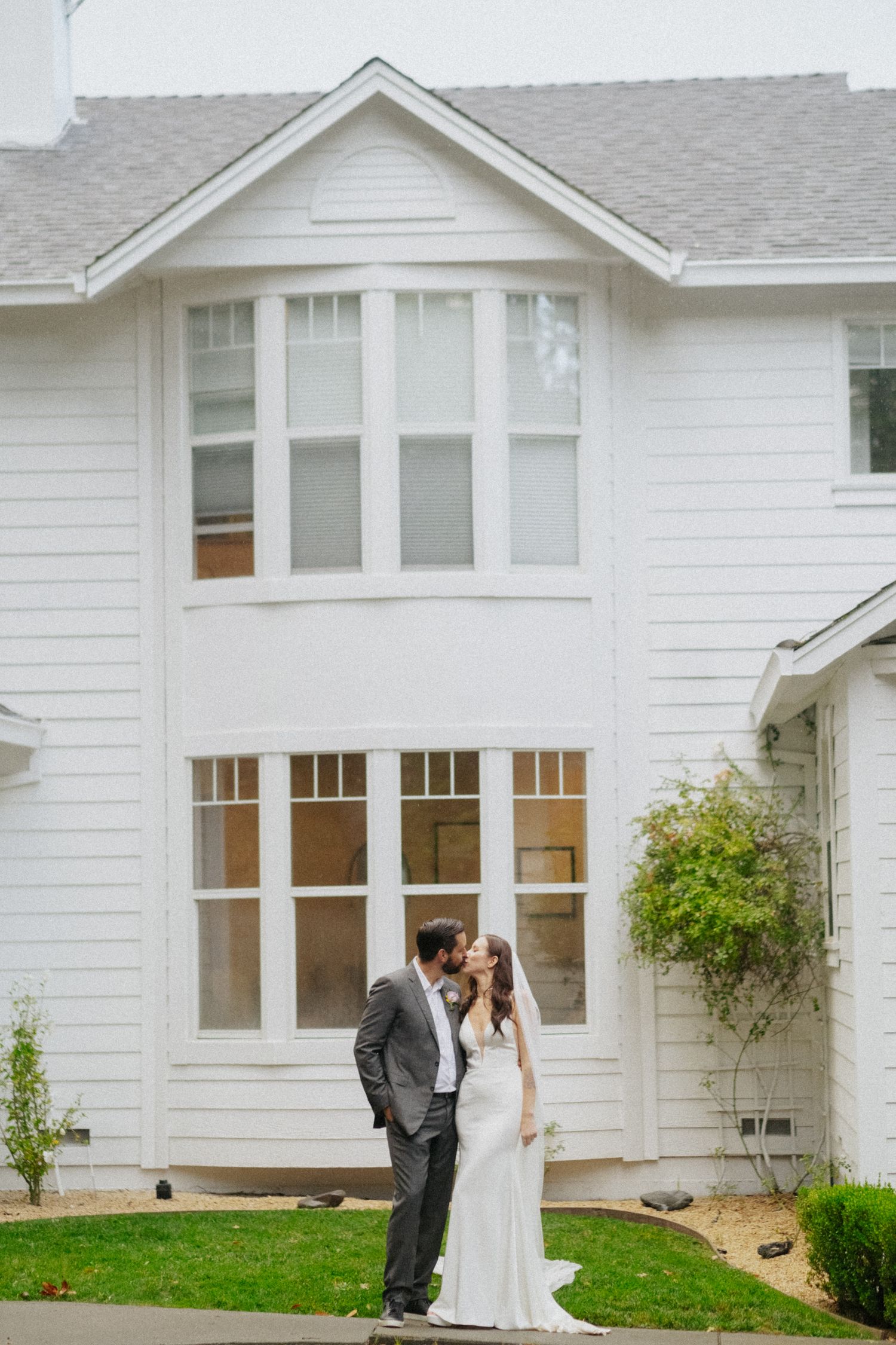 Bride and groom share a kiss outside a charming white farmhouse on their wedding day.