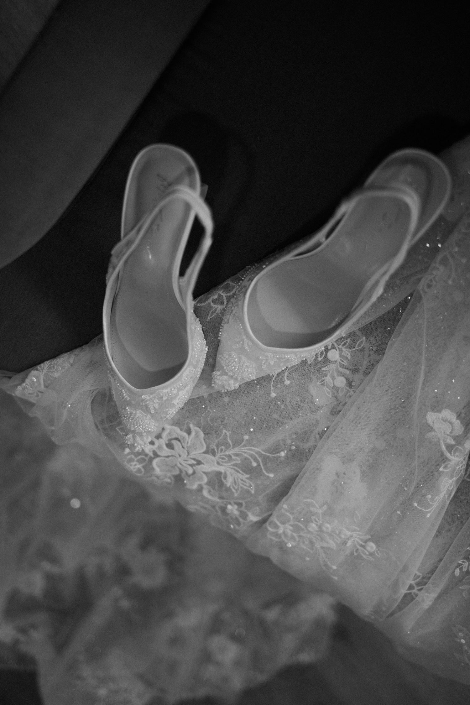 Black and white photo of elegant white lace bridal heels resting on a wedding gown.