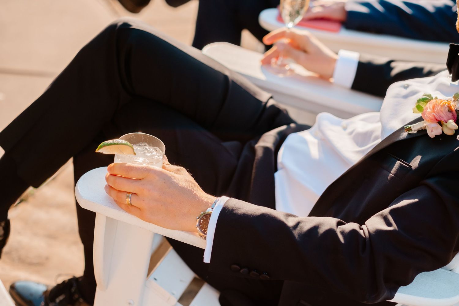 Groom in black tuxedo holding a cocktail glass with ice at an outdoor wedding reception.