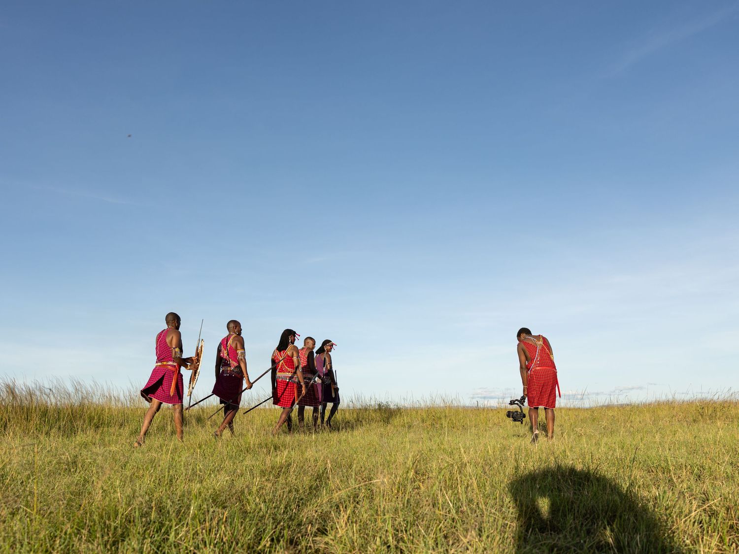 Maasai warriors in red robes stand in tall green grass under a clear blue sky in the African savanna.