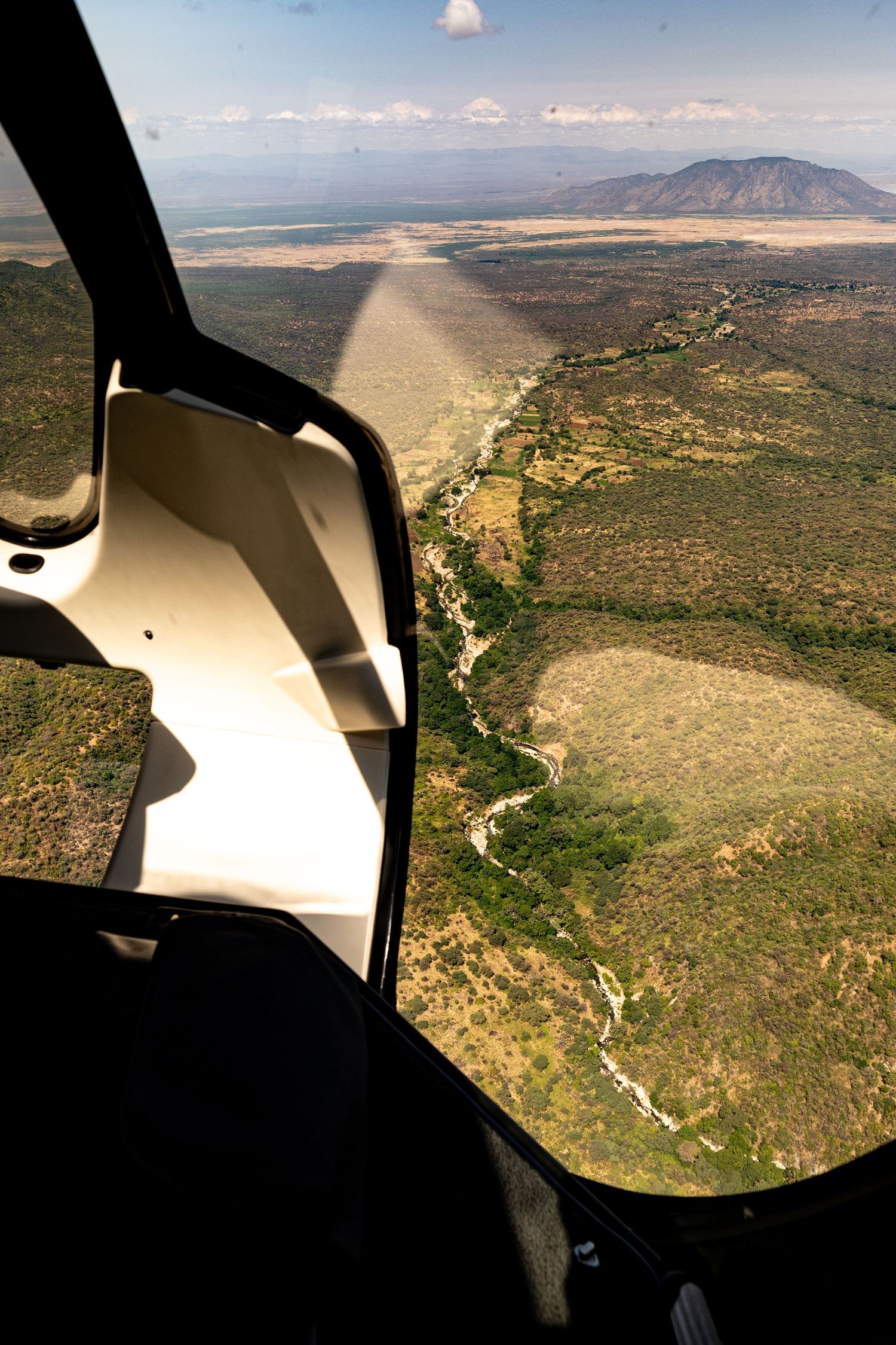 Aerial view from helicopter over African savanna landscape with winding river, dense bush, and mountain in background.