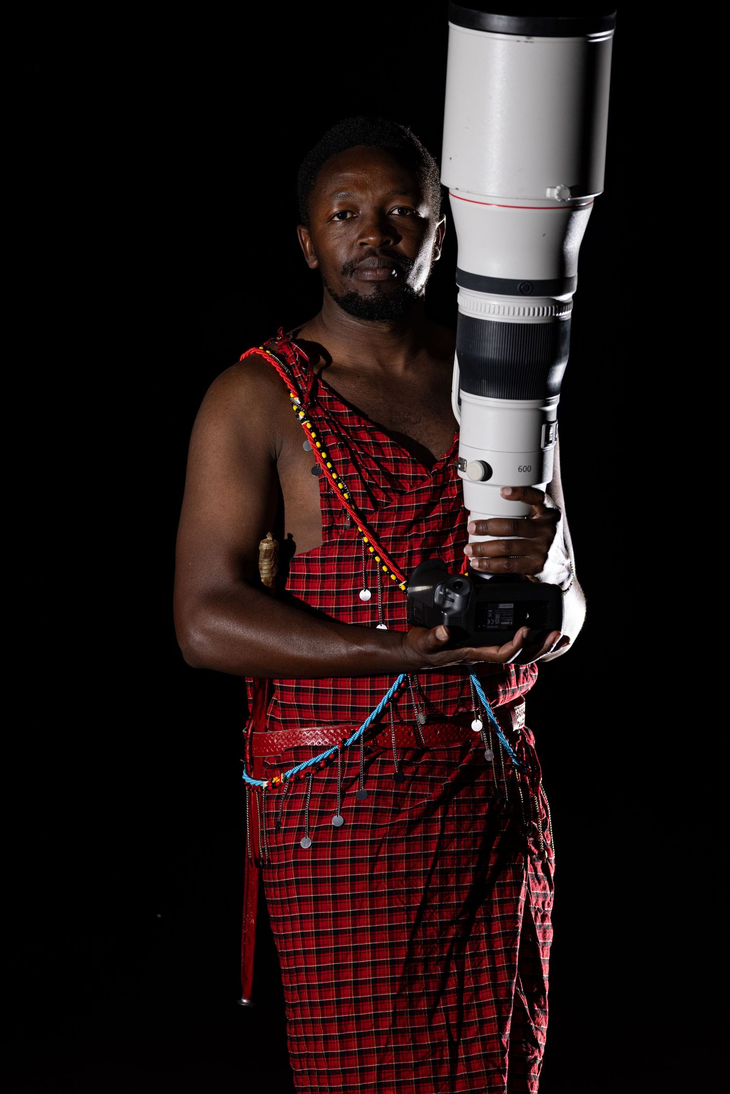A person in a red checkered garment holds and adjusts a large white camera lens against a black background.