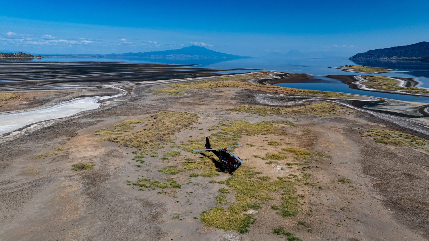 Aerial view of a black helicopter landed on arid terrain near a lake shore with a mountain in the background.