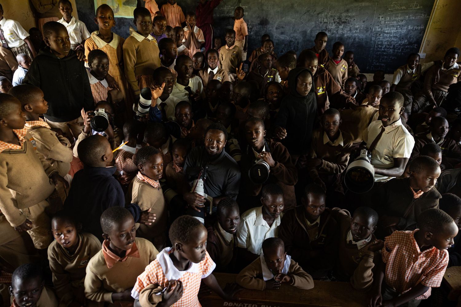 Crowded classroom of African children sitting together in a rustic school room with a chalkboard in background.