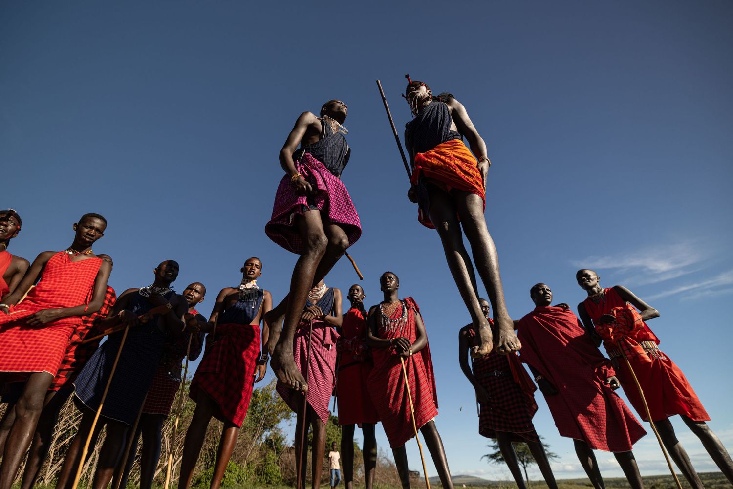 Maasai warriors in red shukas performing traditional jumping dance against blue sky in Kenya.