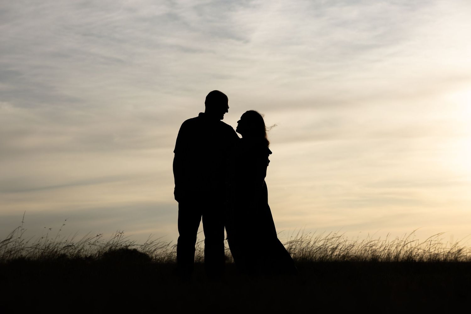 Silhouetted couple standing together against a cloudy sunset sky in a grassy field.