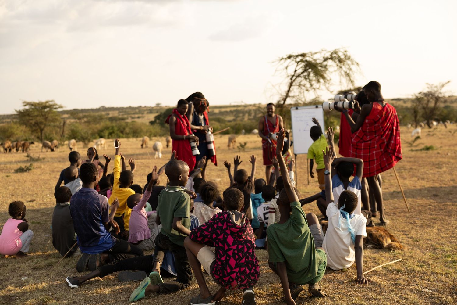 Maasai teacher leads outdoor classroom lesson for children sitting on ground in African savanna landscape.