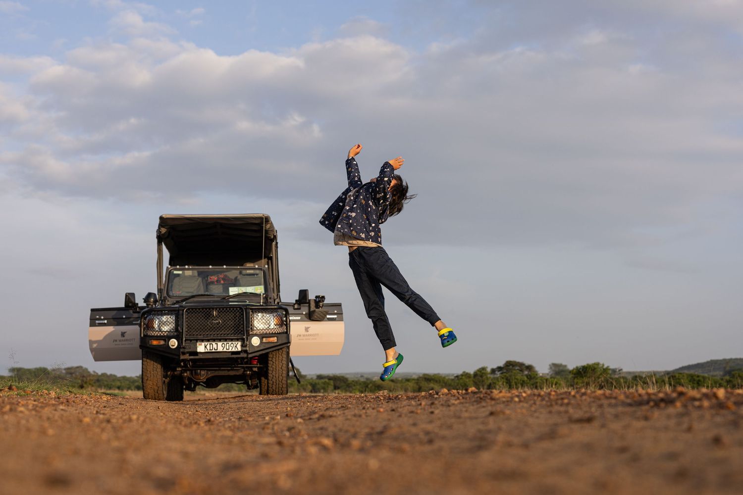 Person jumping joyfully next to a safari jeep on a dirt road under a cloudy sky in Africa.