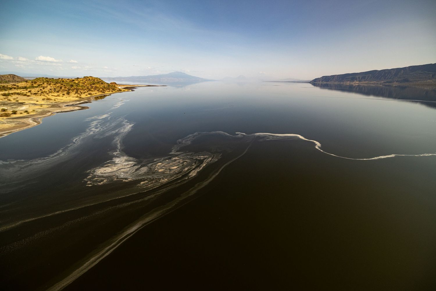 Aerial view of a vast calm lake with dark water, sandy shores, and distant mountains under a clear blue sky.