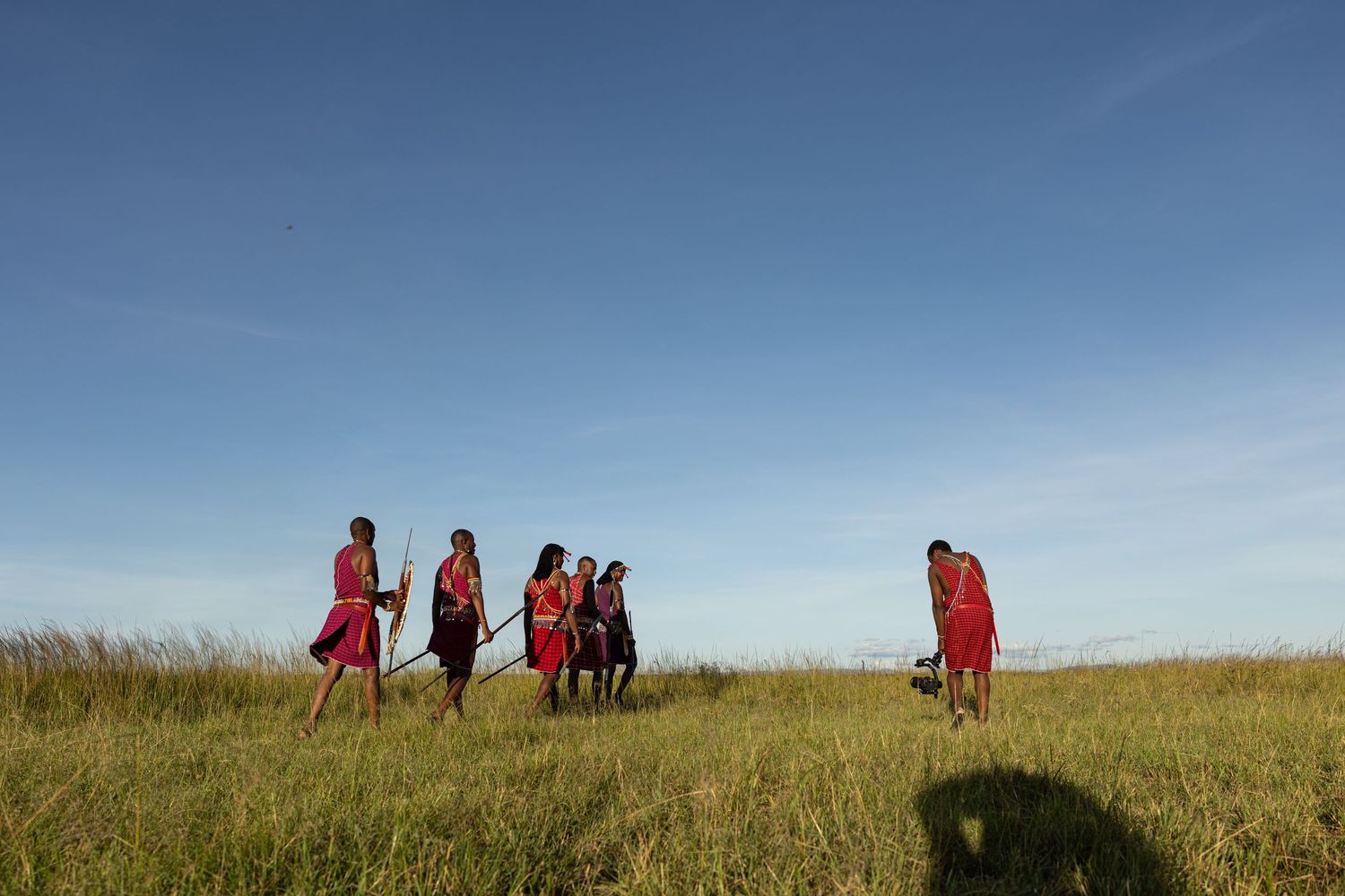 Maasai warriors in red shukas walk through tall grass savanna under a clear blue sky in Africa.