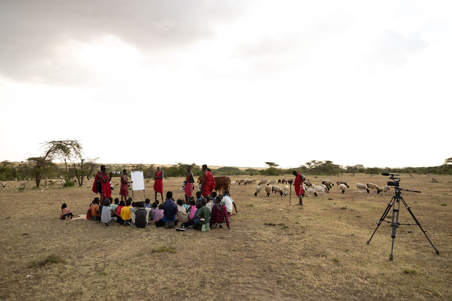 Maasai children gather in a circle outdoors on arid land for an open-air lesson, with a camera tripod nearby.