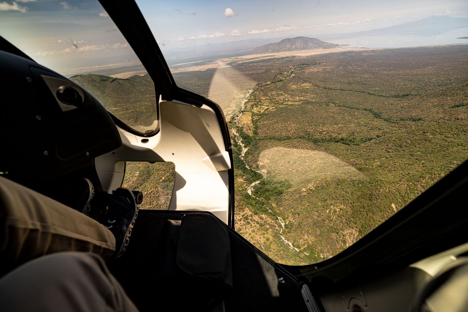 Aerial view from helicopter cockpit over vast African savanna landscape with mountains and coastline in the distance.