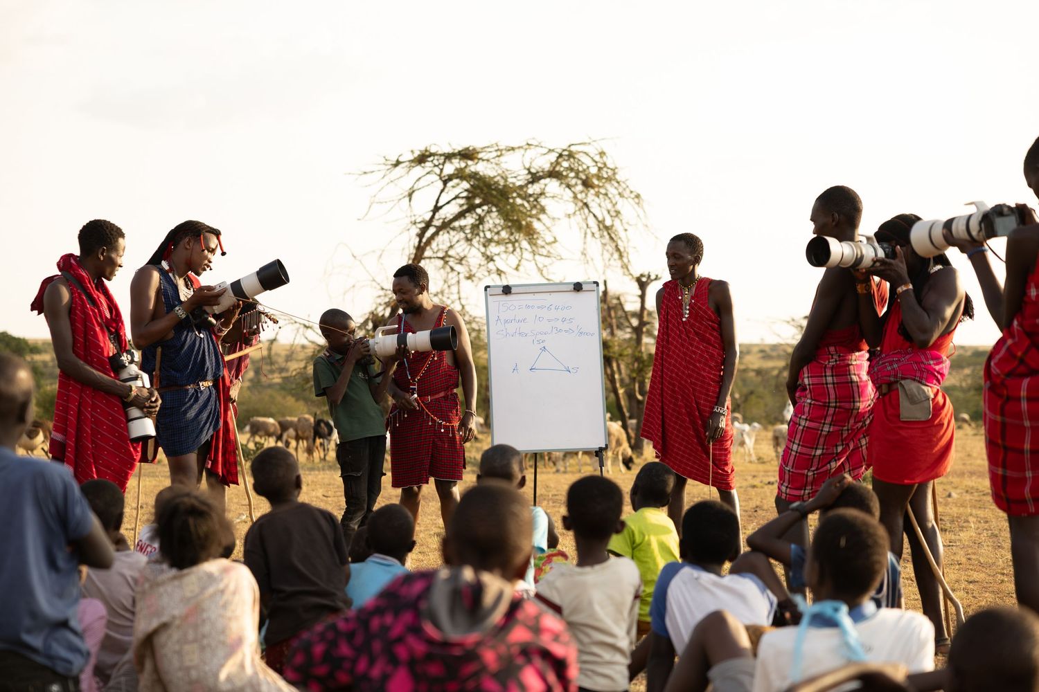 Maasai community gathering outdoors with photographers and whiteboard presentation in African savanna setting.