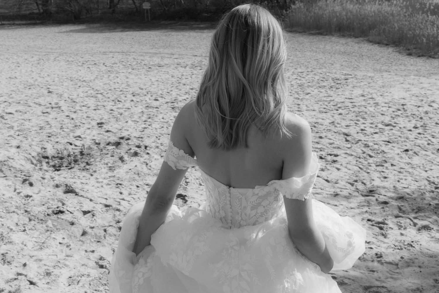 Black and white photo of a bride in an off-shoulder wedding gown sitting on sandy beach, viewed from behind.