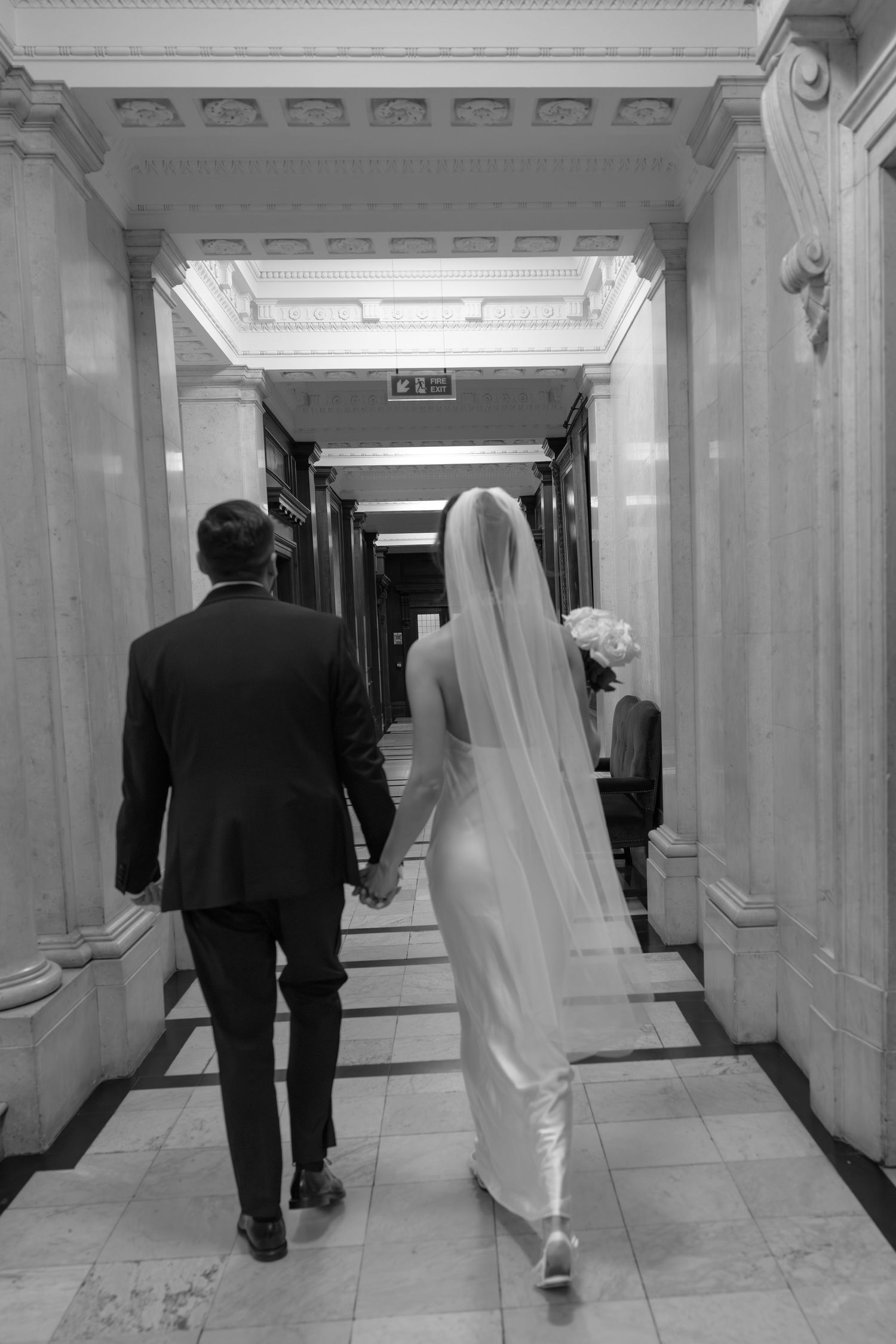 Black and white photo of newlyweds walking hand in hand down elegant marble hallway.