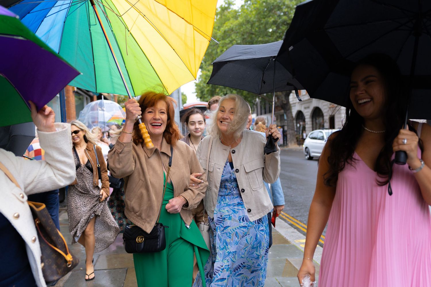 Wedding guests walk under colorful umbrellas in the rain on a London street, smiling and celebrating.
