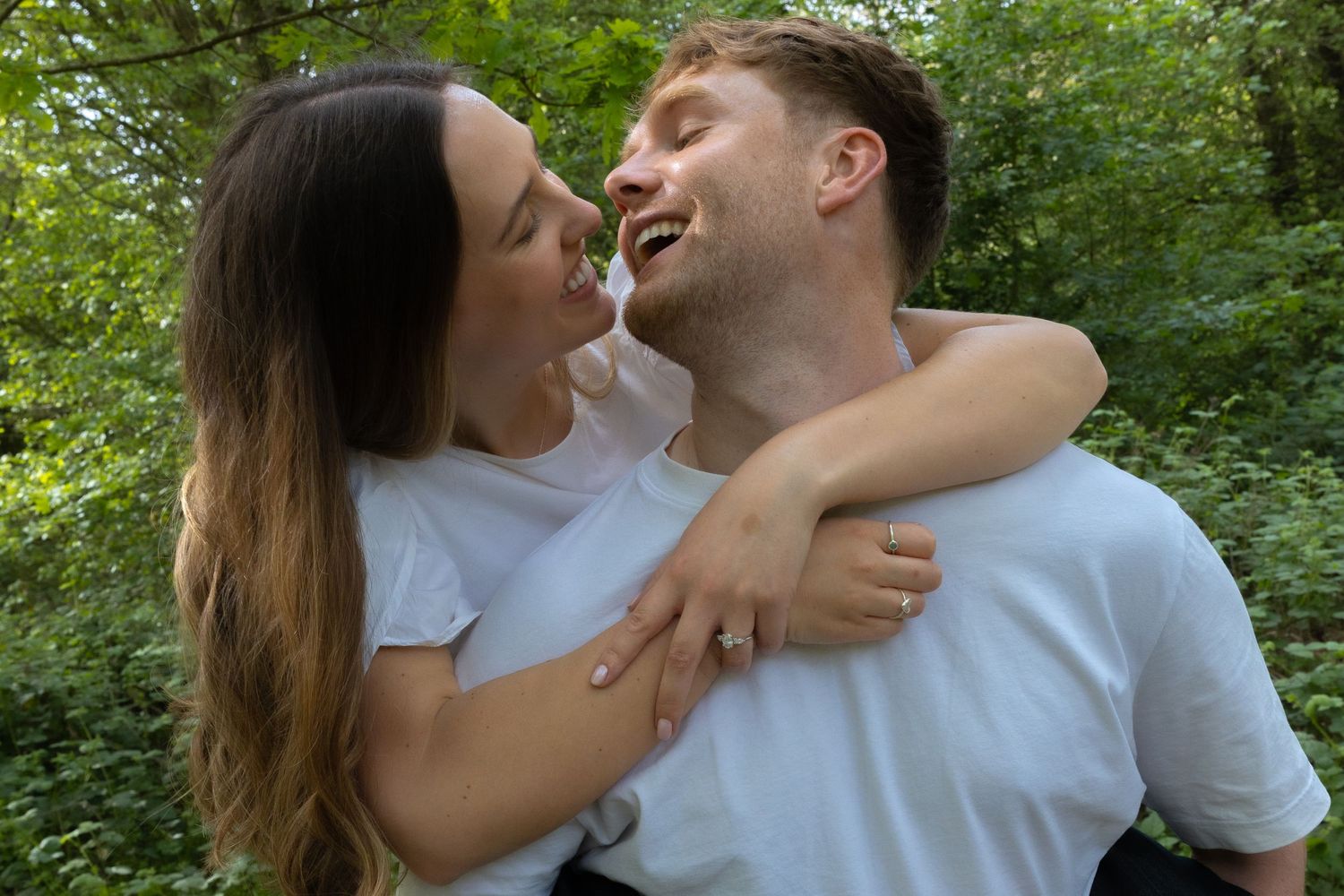 Happy couple laughing and hugging outdoors in a green forest setting, wearing matching white t-shirts.