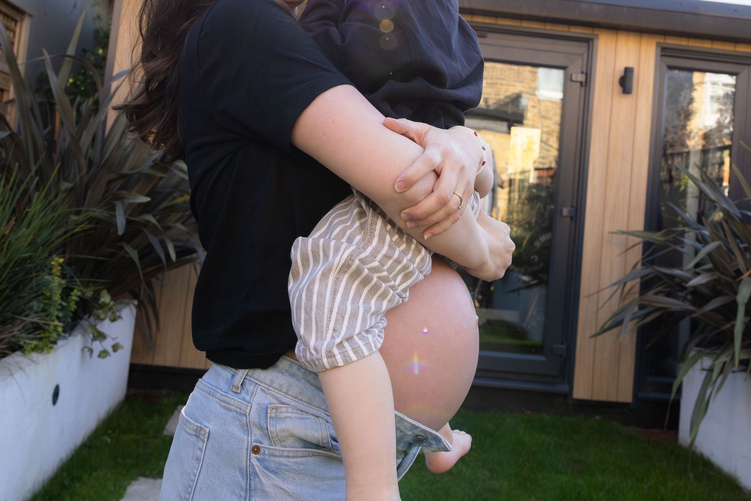 Pregnant woman holding a toddler with stuffed animal in a garden setting.