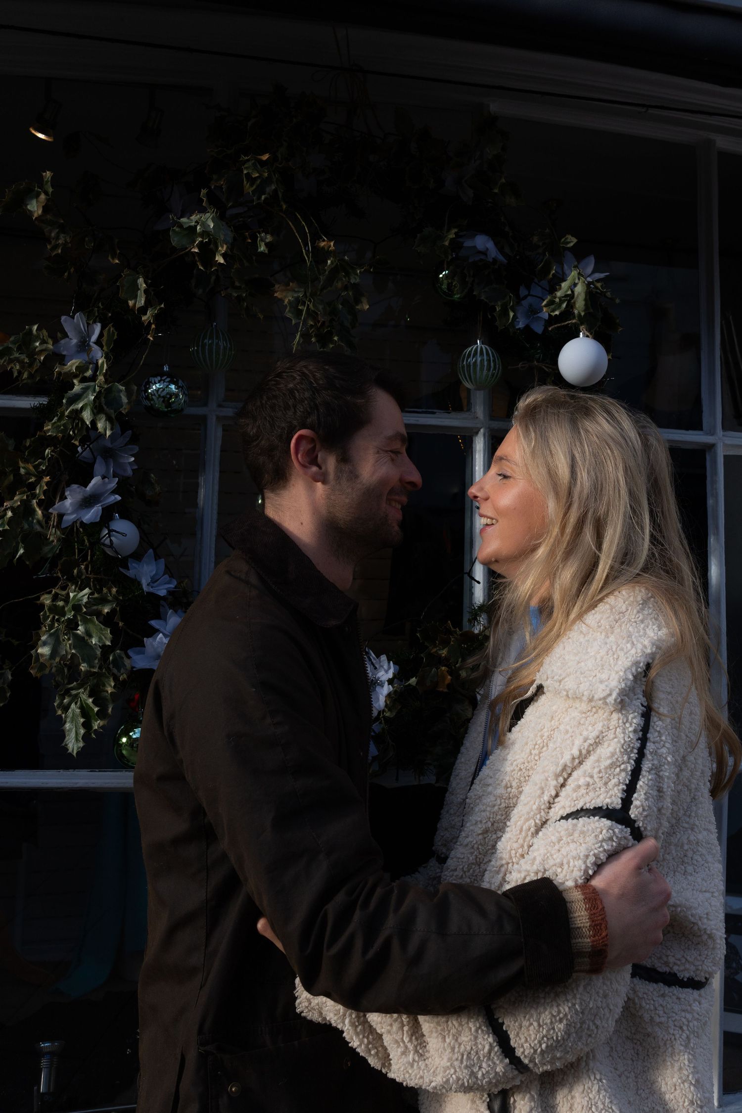 A couple shares an intimate moment near a window decorated with holiday ornaments and greenery.