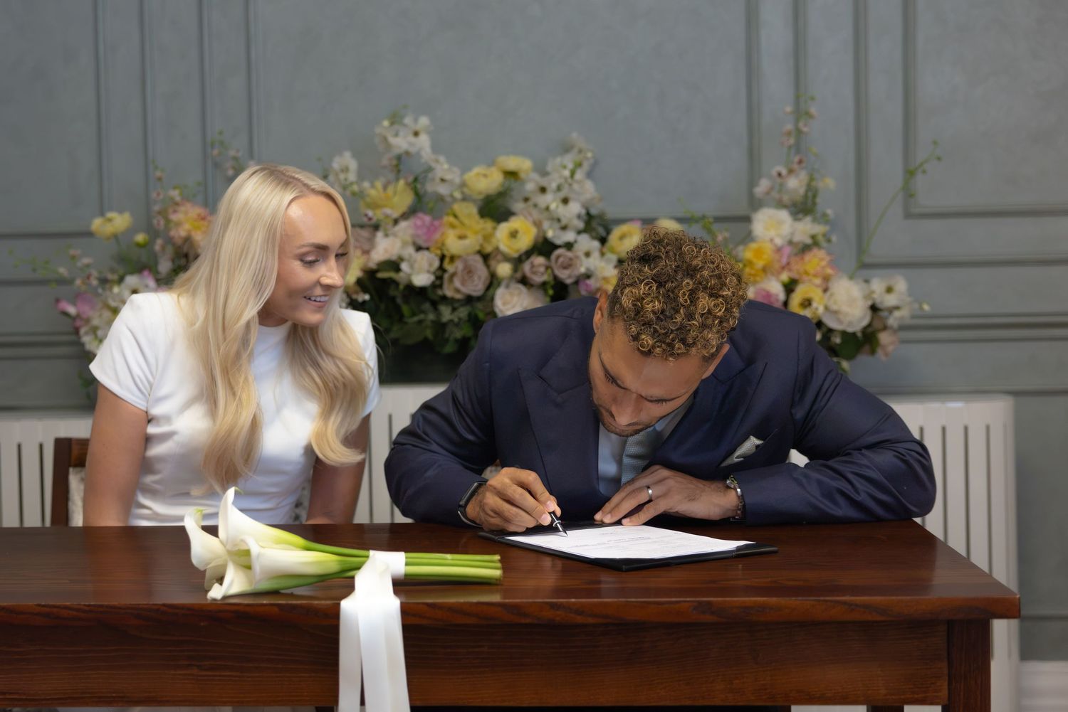 Couple signing documents at a wooden table with floral arrangement, woman in white top watching man in suit write.