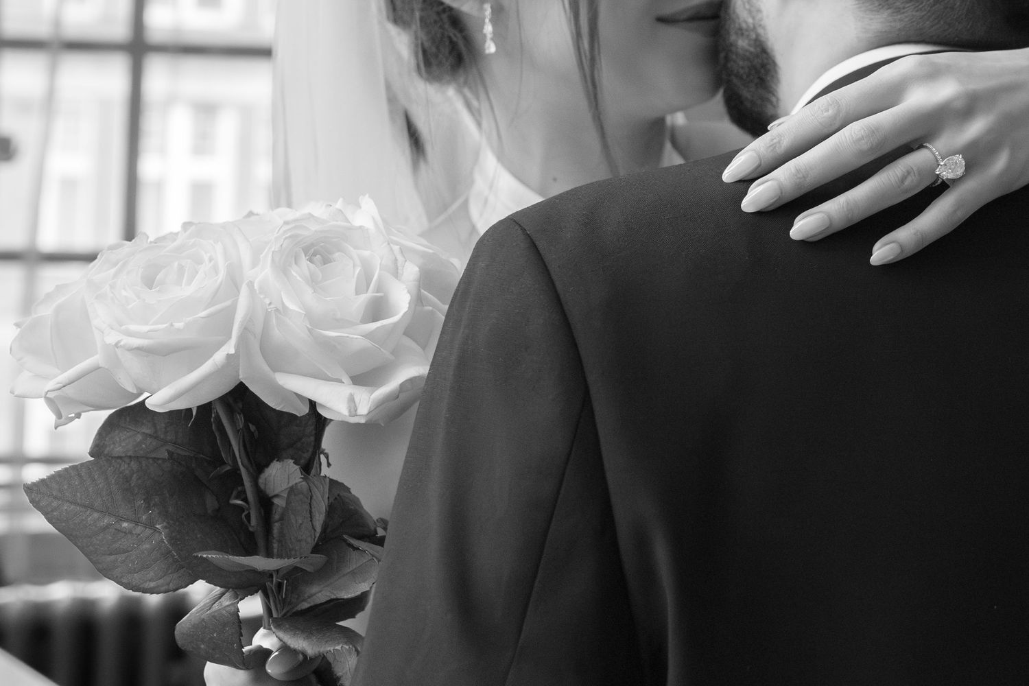 Black and white wedding photo of bride holding white roses, embracing groom, with diamond engagement ring visible.
