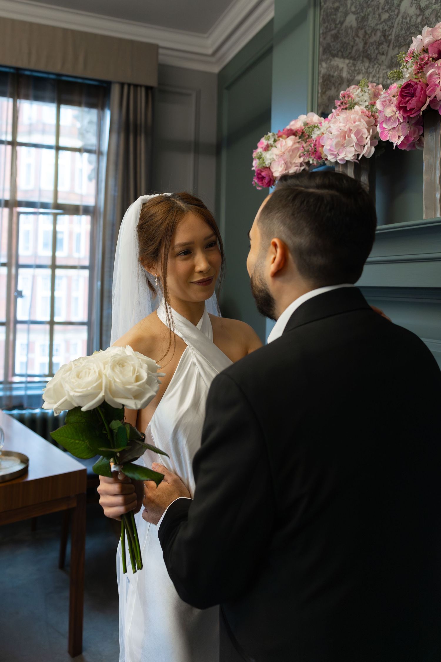 Bride in white halter gown holding white roses faces groom in black suit during indoor wedding ceremony.