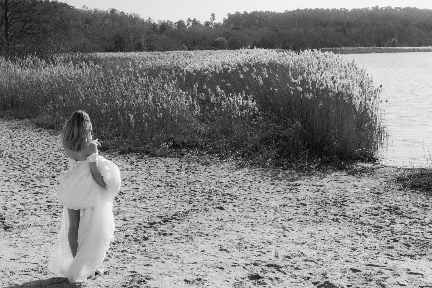 Black and white photo of a woman in a white dress standing on a sandy beach near tall reeds and a calm lake.