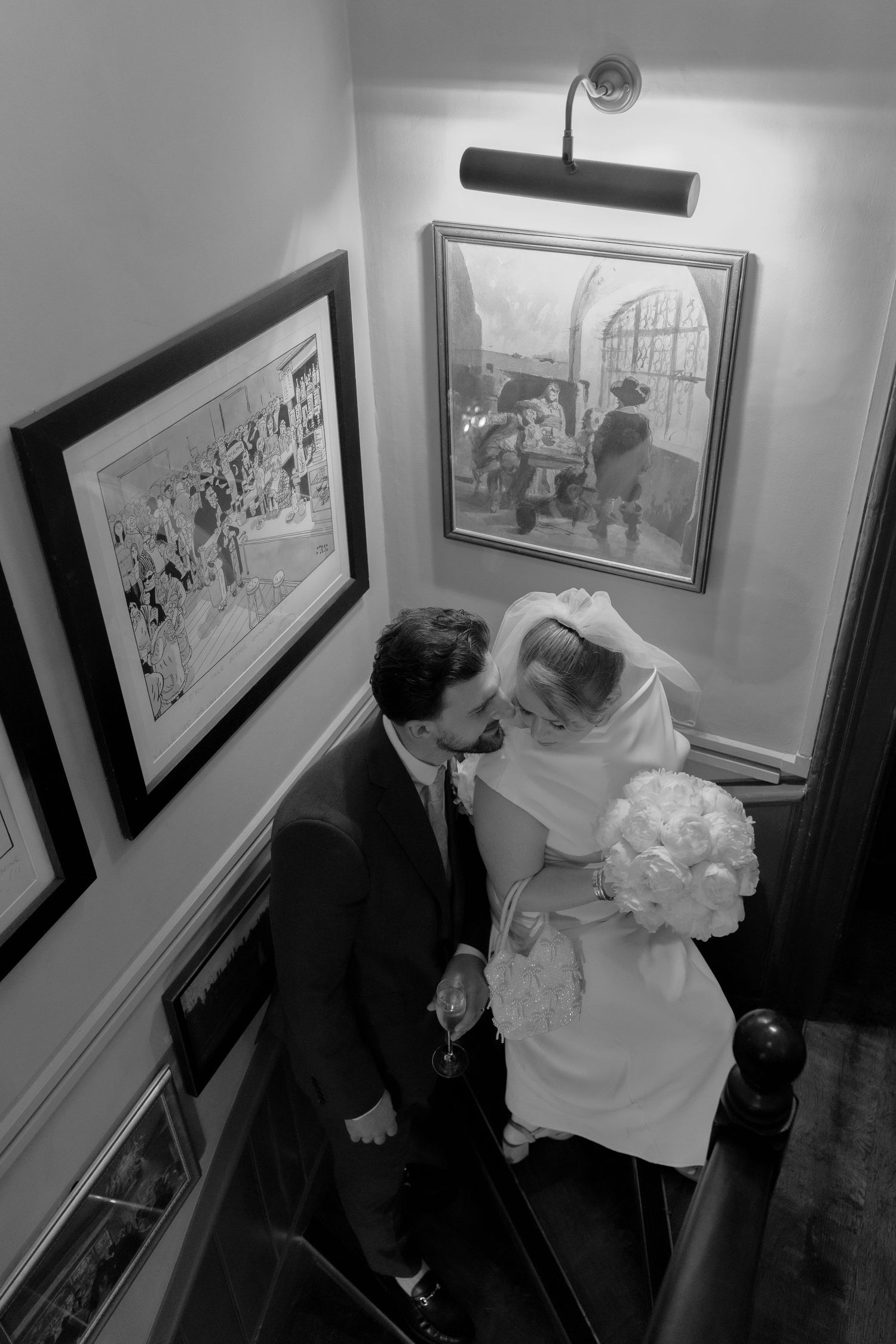 Black and white photo of bride and groom sharing a kiss on stairs surrounded by framed artwork.