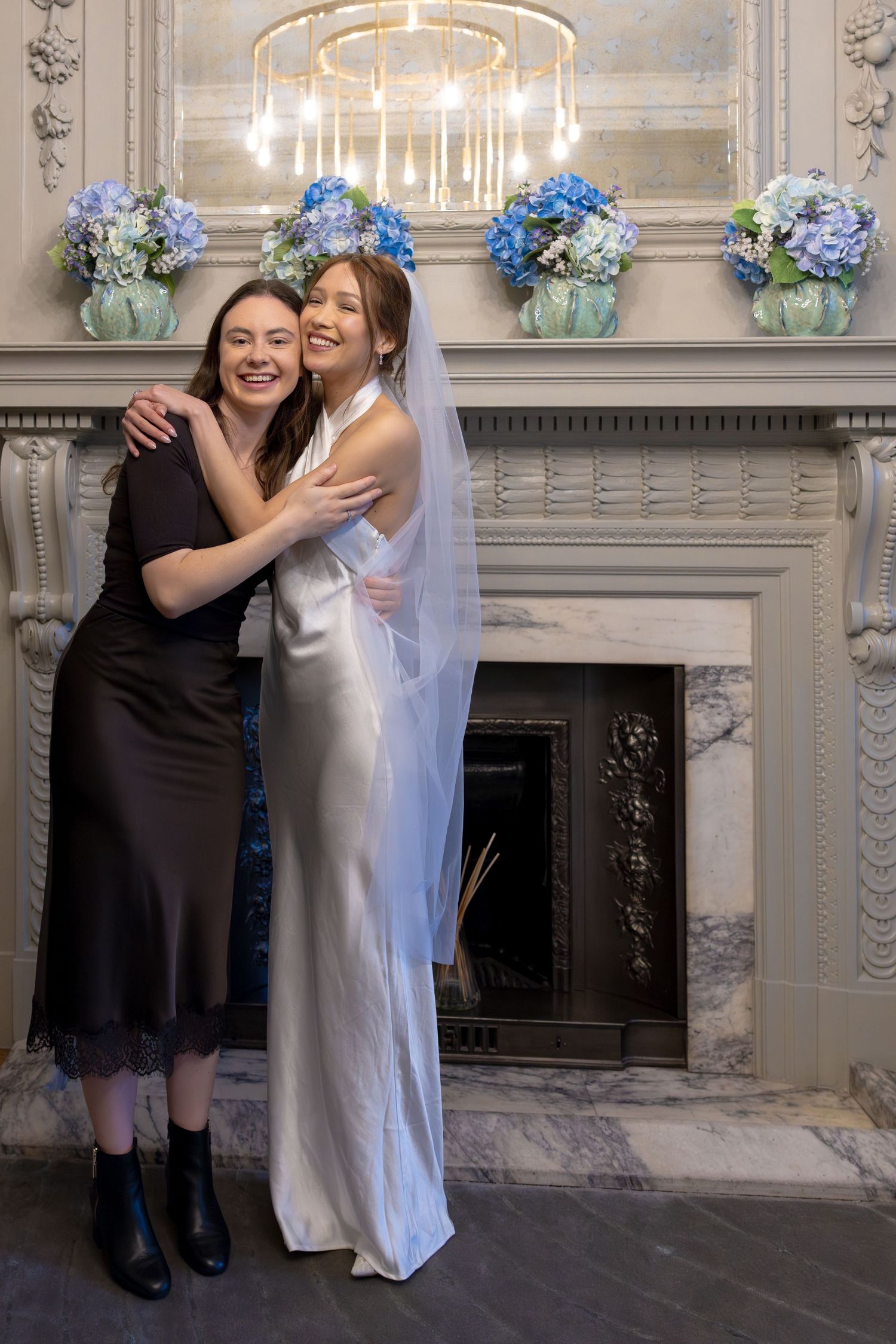 Bride in white satin gown and veil embracing female guest in black dress at wedding venue.