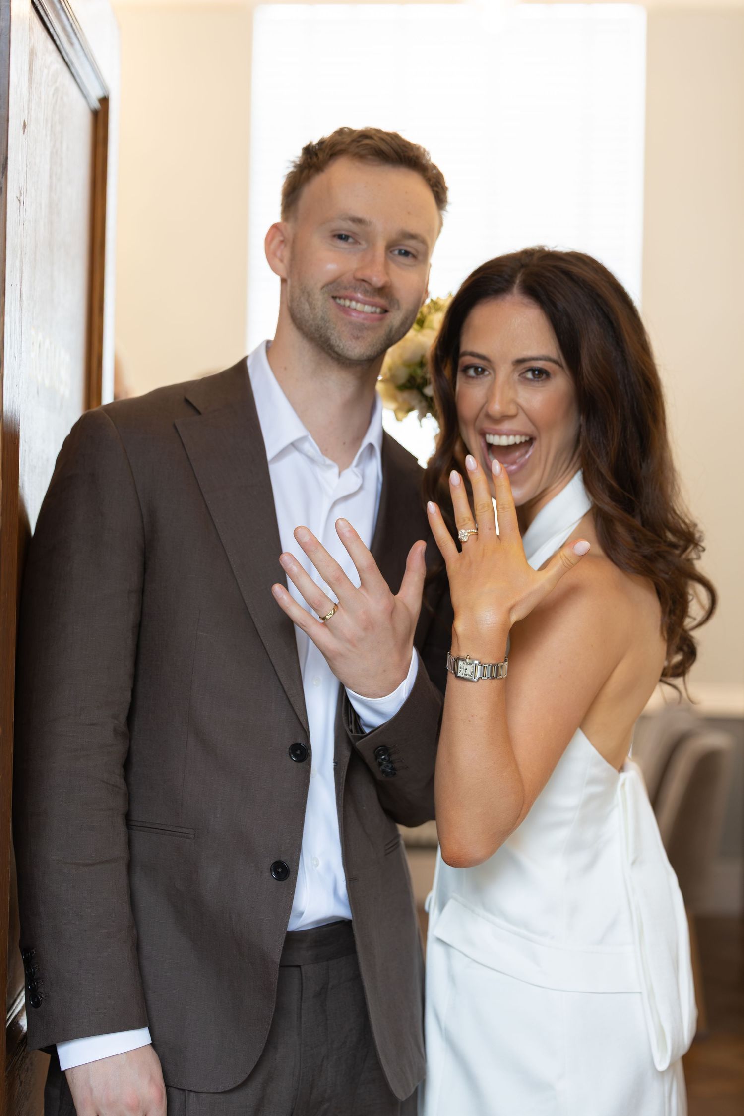 Happy couple showing off wedding rings, woman in white dress and man in brown suit, smiling at camera.
