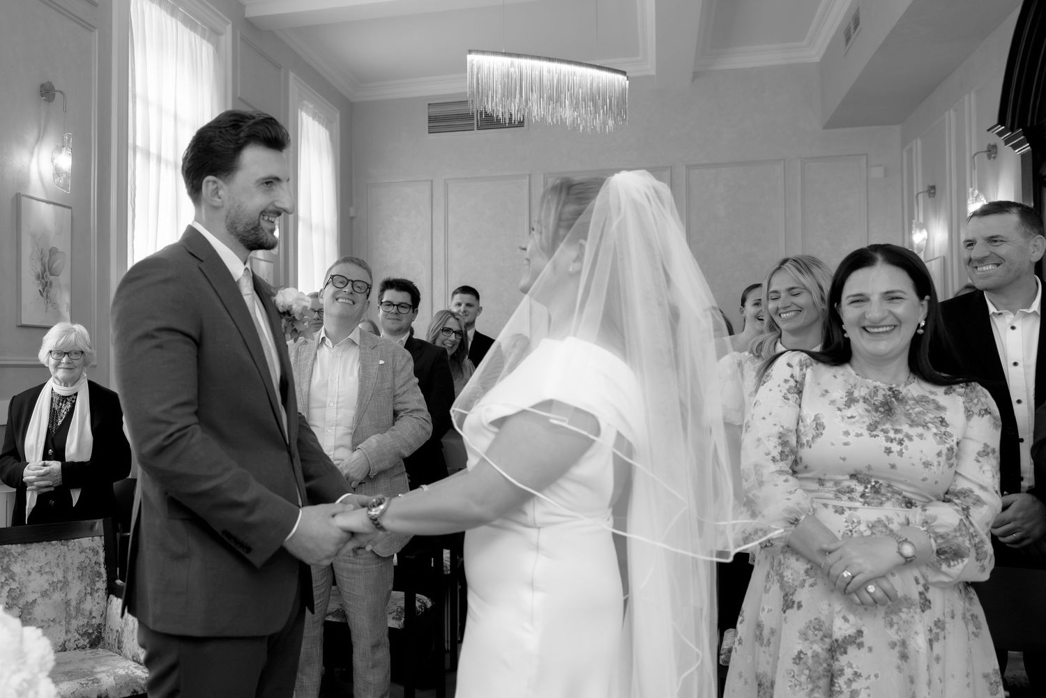 Black and white wedding ceremony photo of bride and groom holding hands at the altar with smiling guests in background.