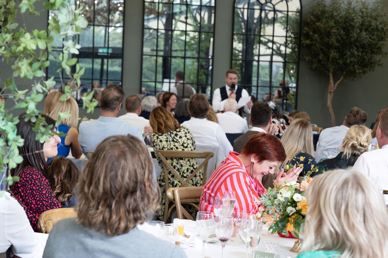 Wedding reception guests seated at round tables in a bright, airy venue with large windows and floral centerpieces.