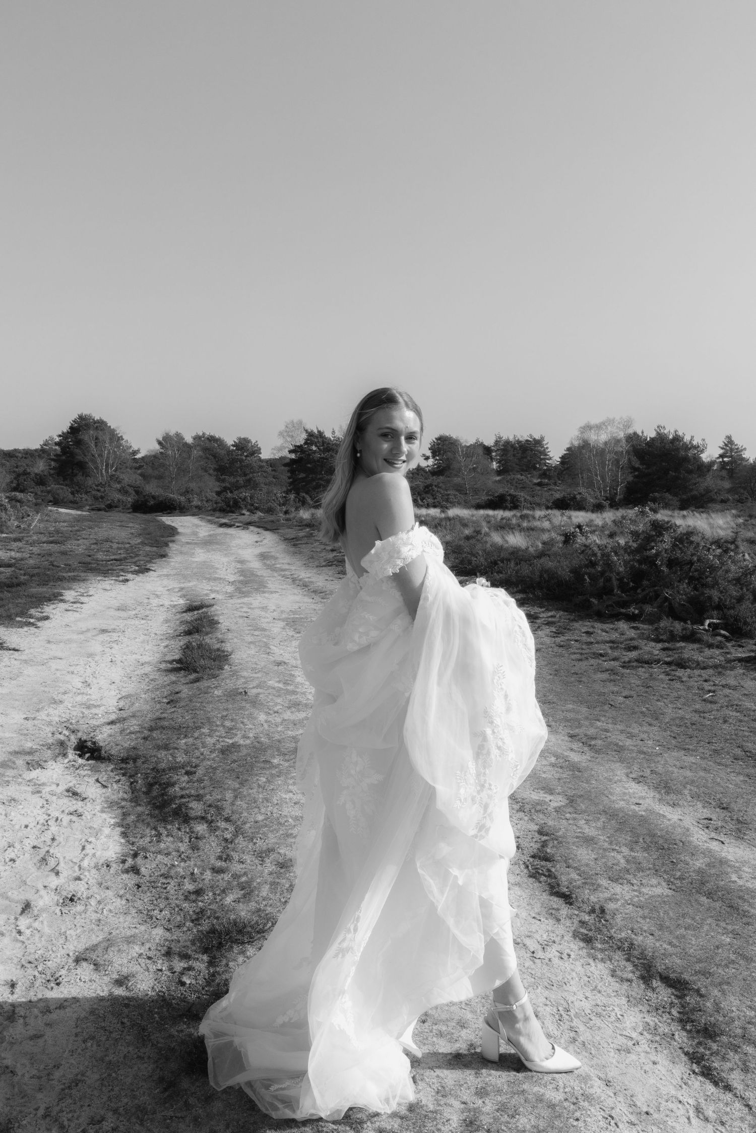 Black and white photo of a bride in a flowing white gown standing on a dirt path surrounded by nature.