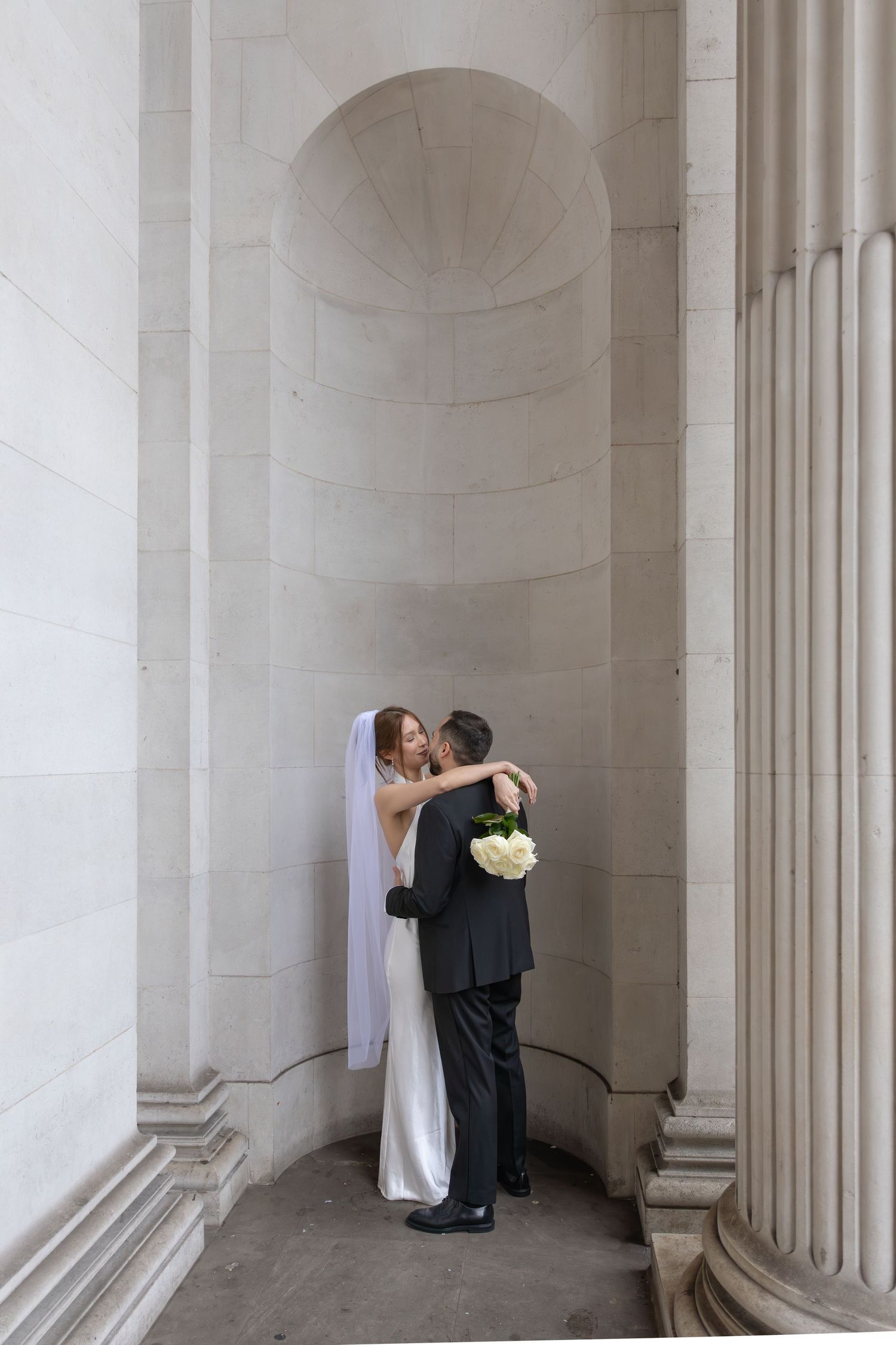 Bride and groom sharing a kiss between grand stone columns at a classical wedding venue.