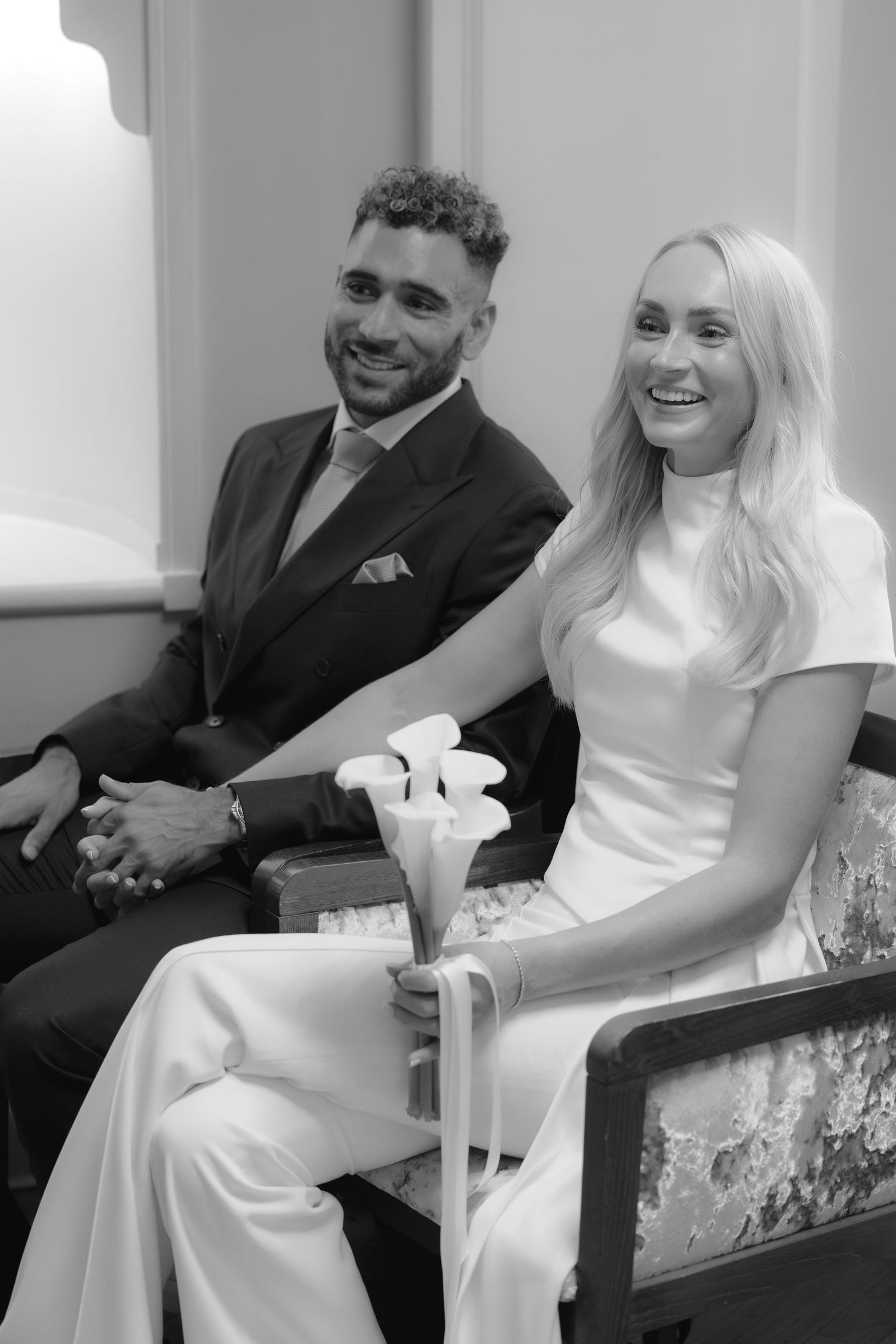 Black and white photo of a smiling bride holding calla lilies seated beside a groom in a suit at a wedding ceremony.