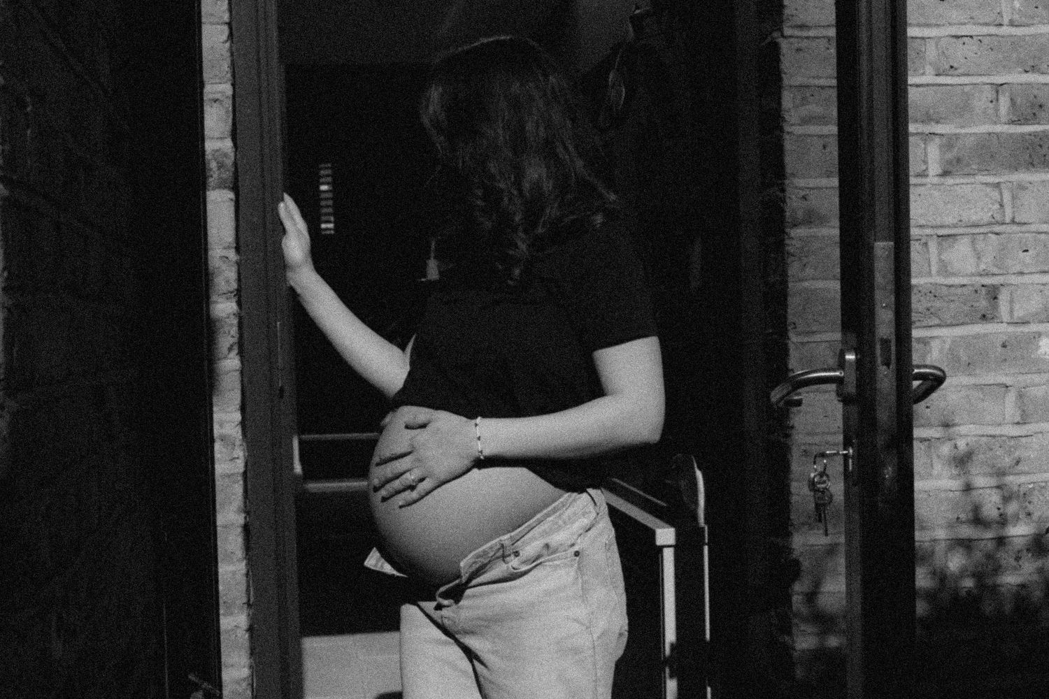 Black and white photo of pregnant woman cradling baby bump while standing in doorway of brick building.