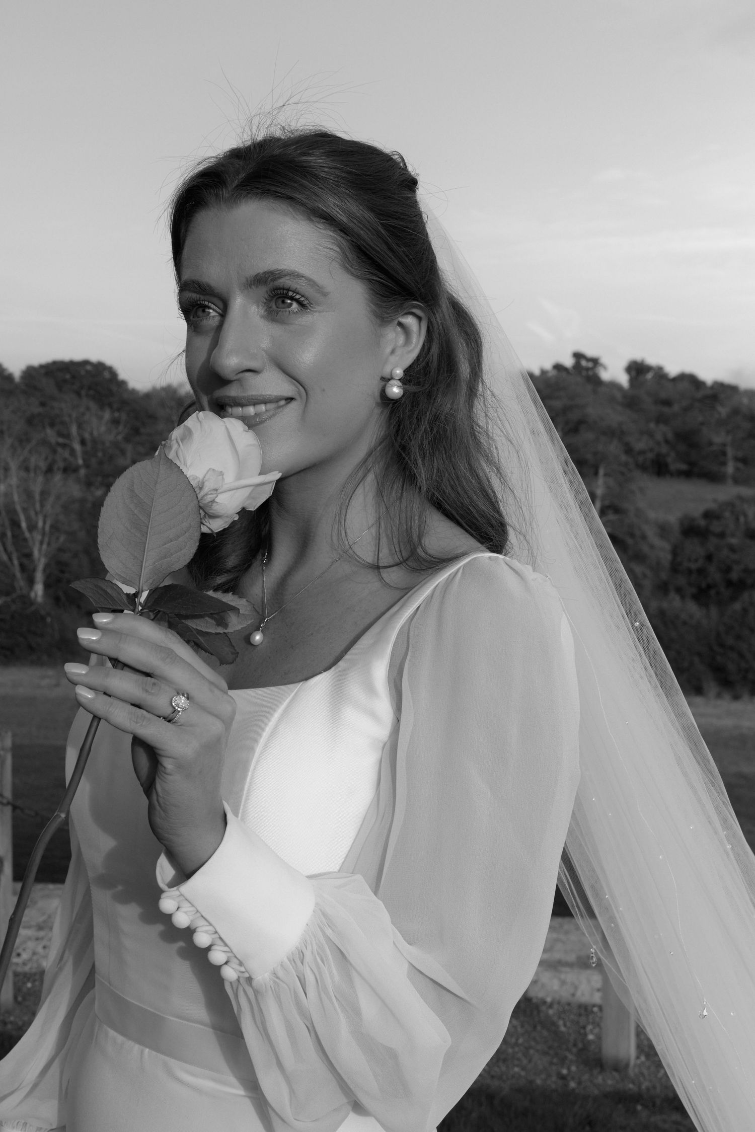 A black and white portrait of a bride smelling a rose while wearing a flowing veil.