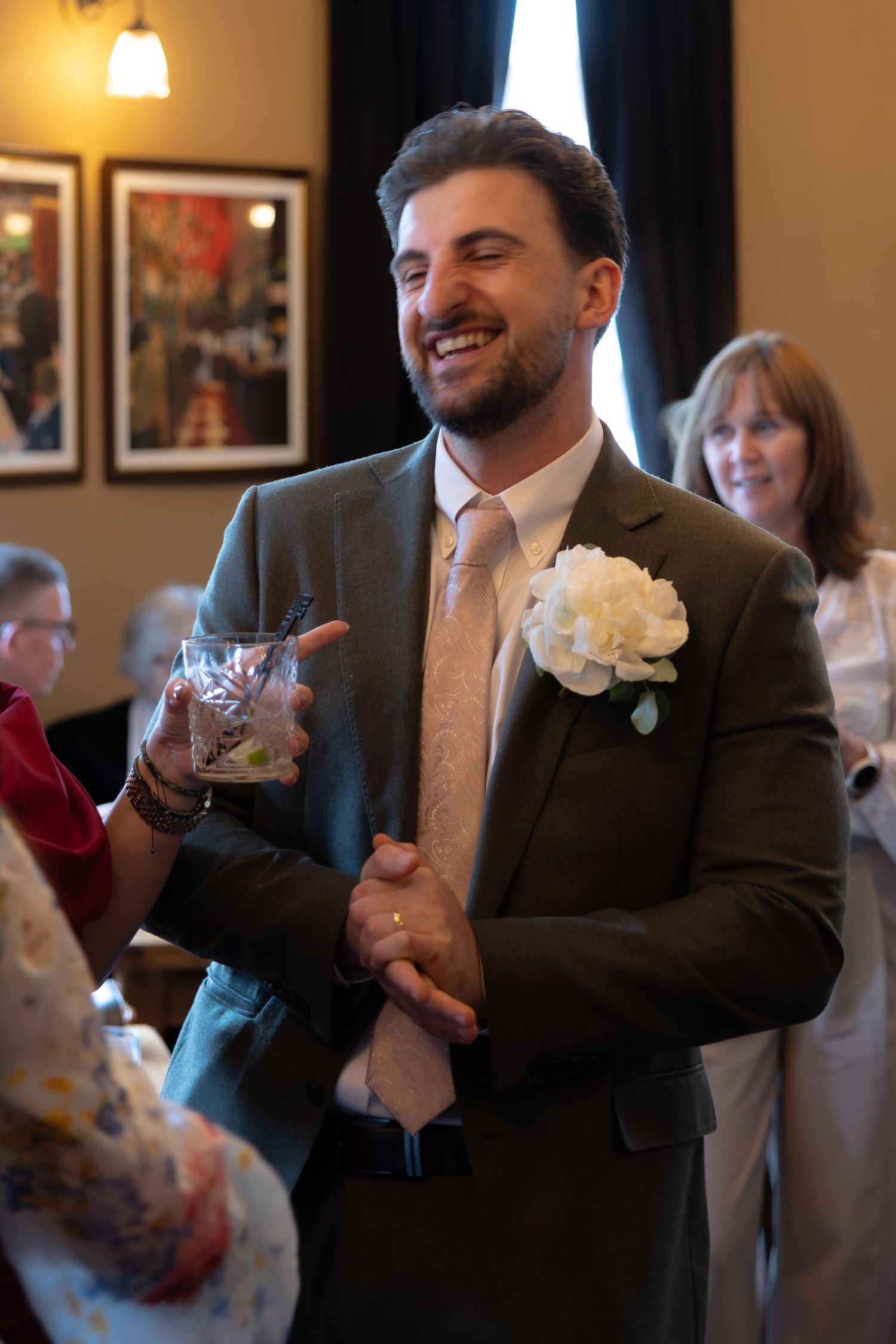 Smiling groom in gray suit with white boutonniere holding a drink at wedding reception.