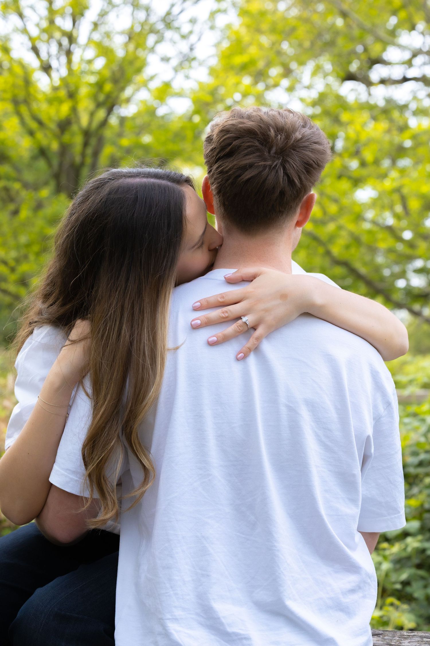 A couple shares a tender kiss outdoors, with the woman embracing the man from behind amid lush green trees.