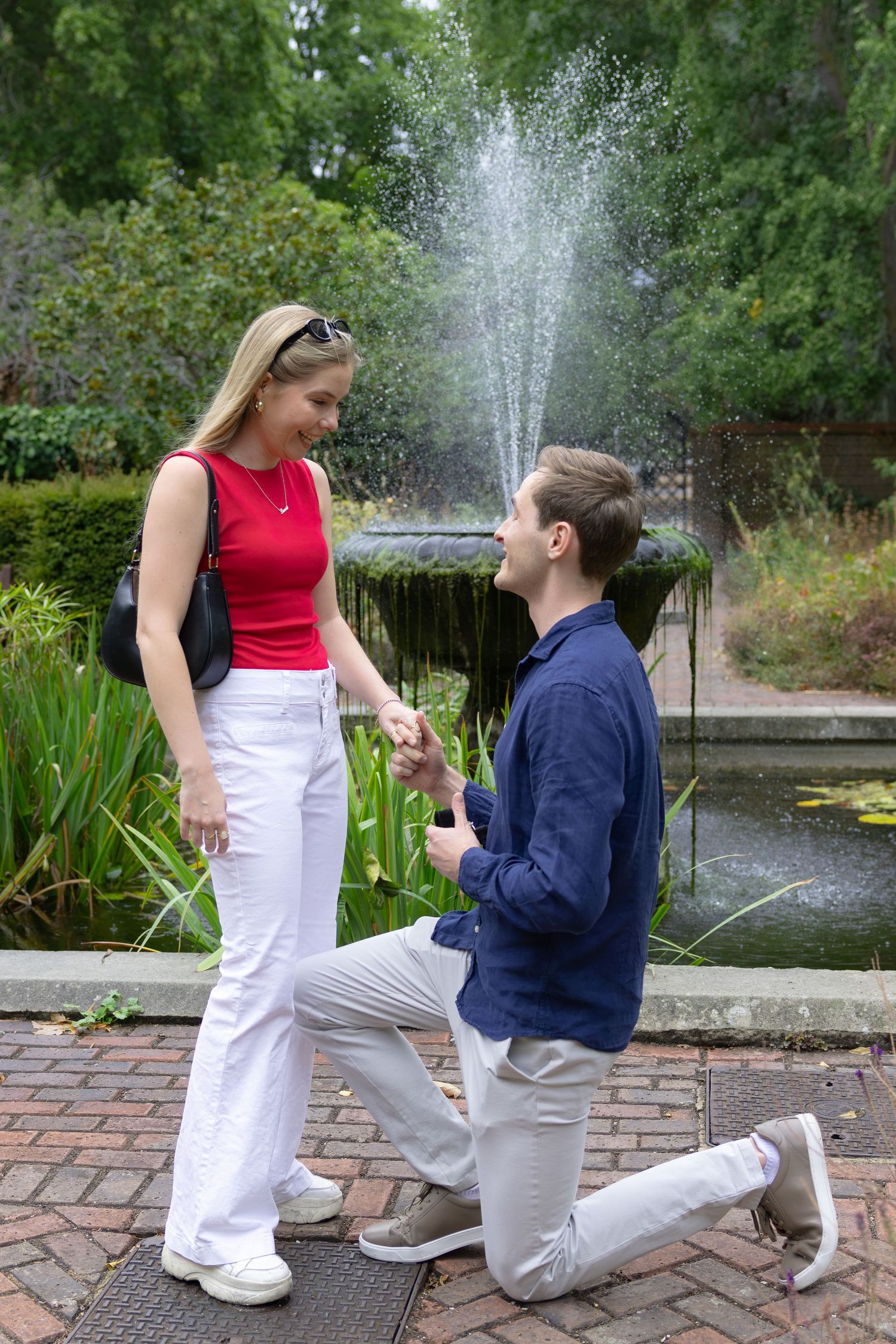 Man proposes on one knee to woman near a garden fountain, holding a ring box while she smiles.