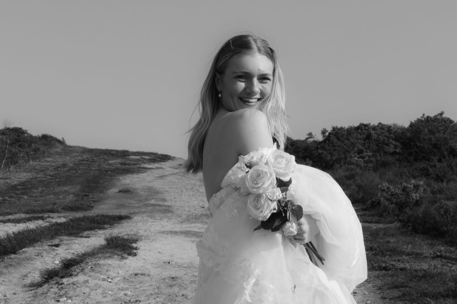 Black and white photo of a smiling bride in a white gown holding a bouquet on a scenic dirt path.