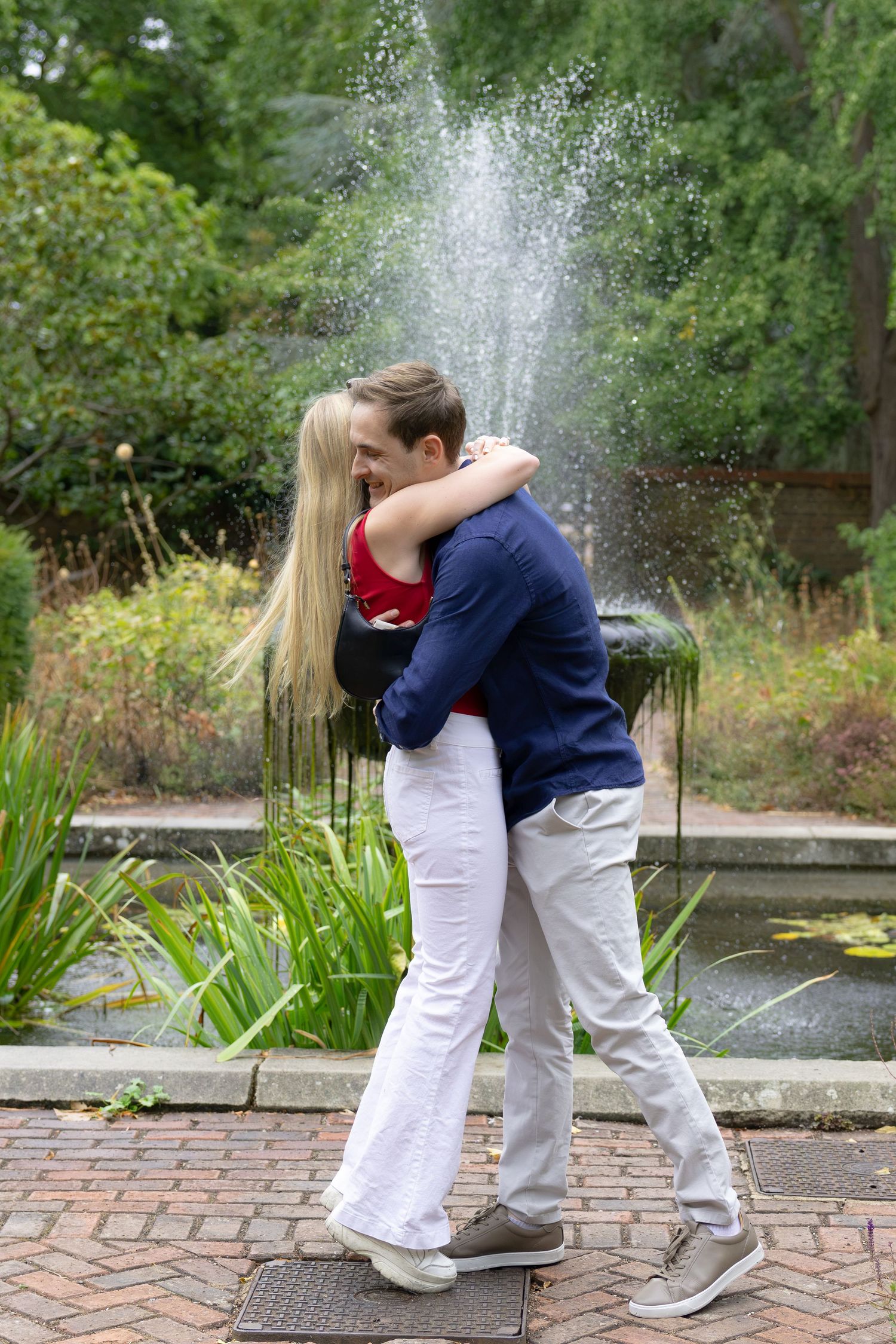 Couple embracing romantically in front of a garden fountain surrounded by lush greenery on a brick pathway.