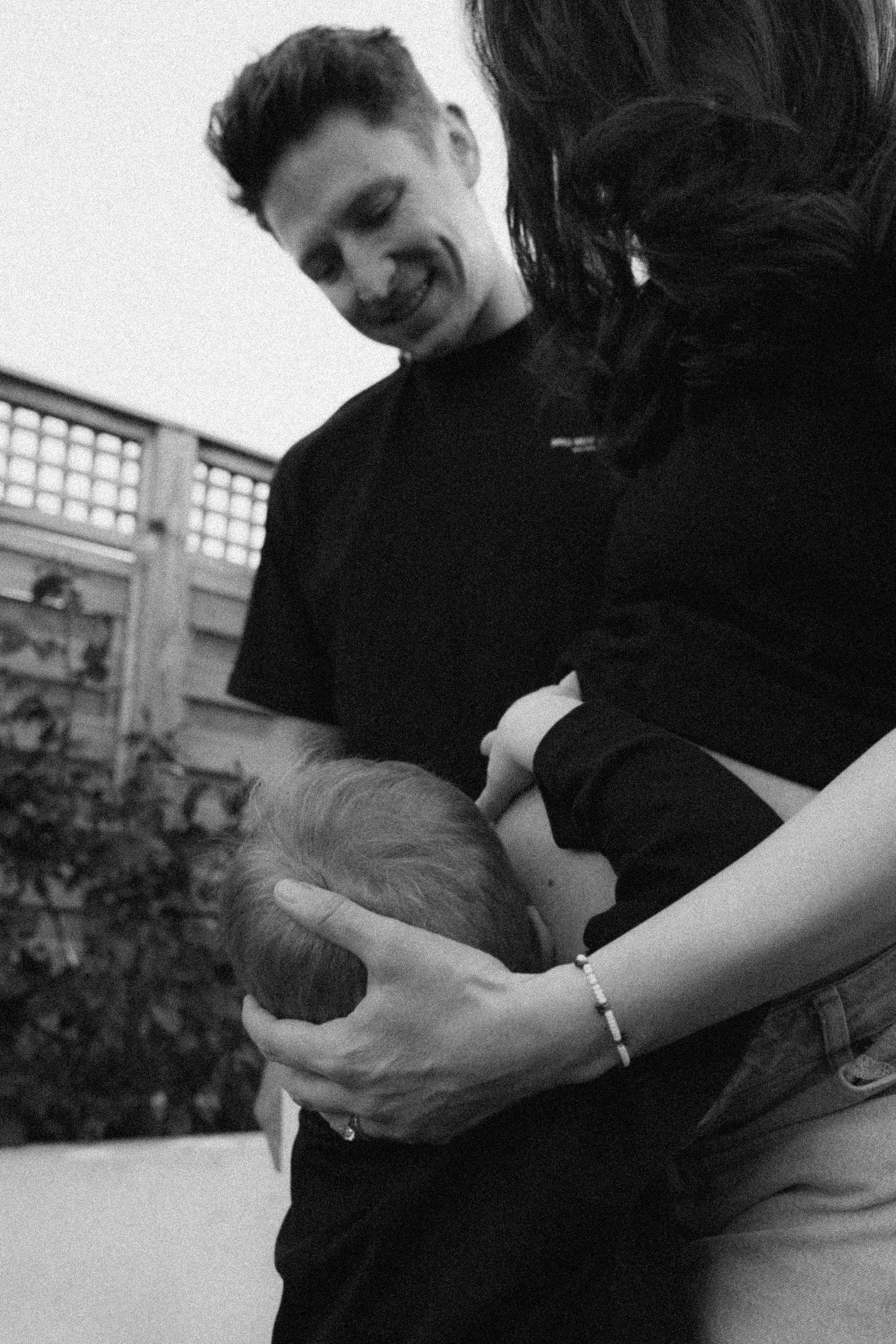 A smiling man in black shirt tenderly holds a newborn baby with a woman in a black and white outdoor photo.