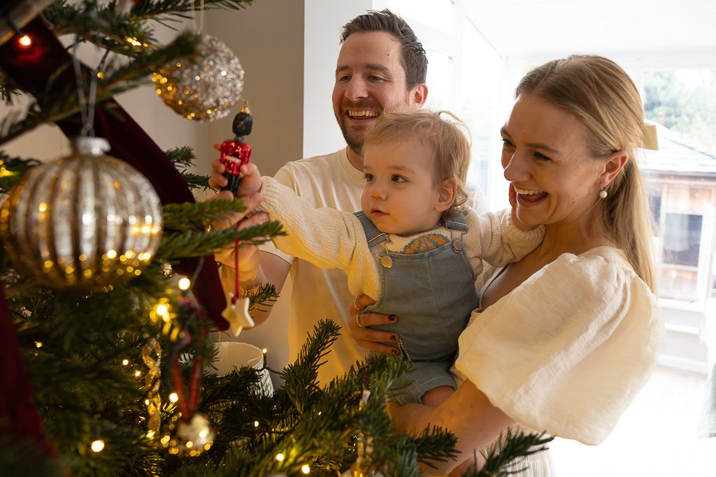 A family decorates their Christmas tree together, reaching for a large silver ornament.