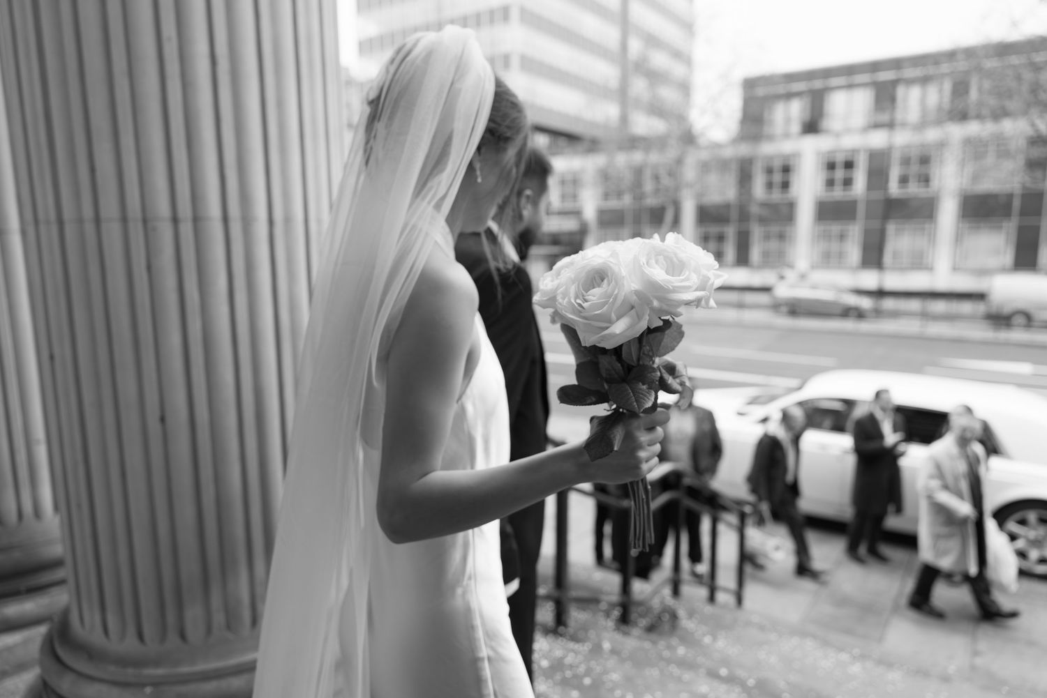 Bride in white gown and veil holds white roses bouquet near ornate column outside wedding venue.