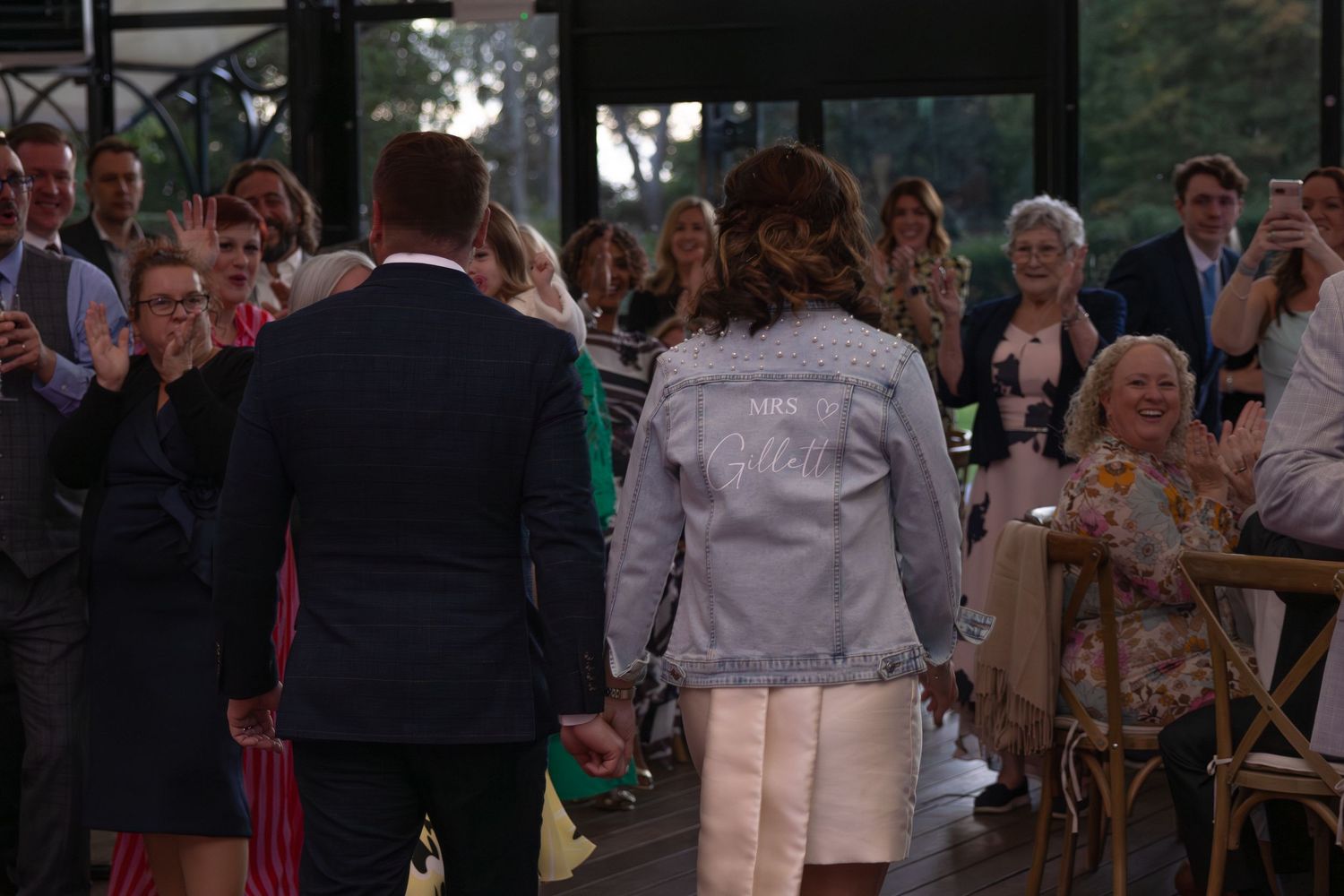 Wedding guests applaud during an outdoor ceremony as a couple stands before them in formal attire.